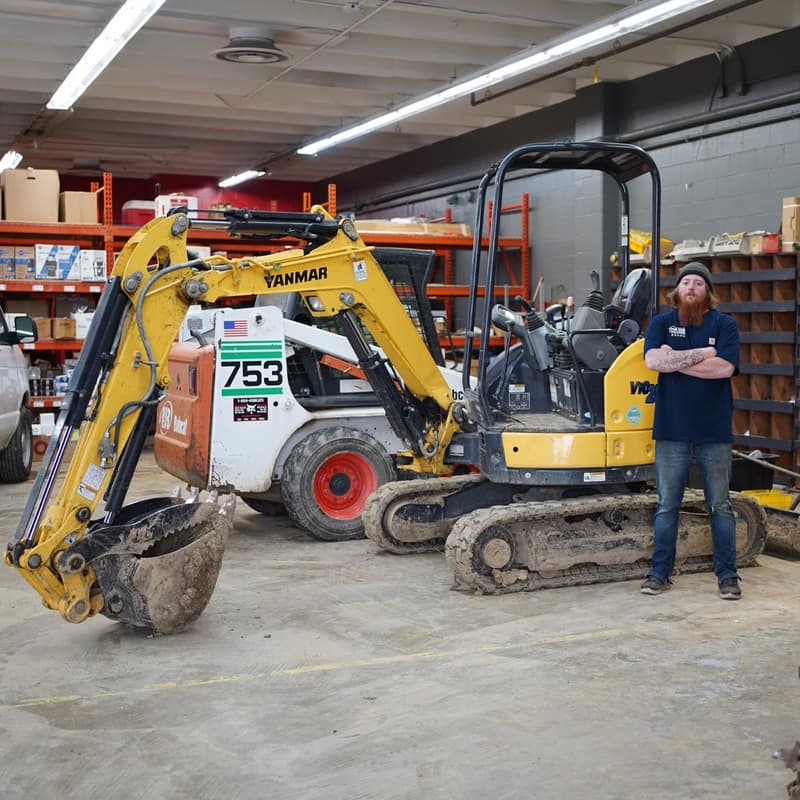 Technician standing beside mini excavator and skid steer in shop